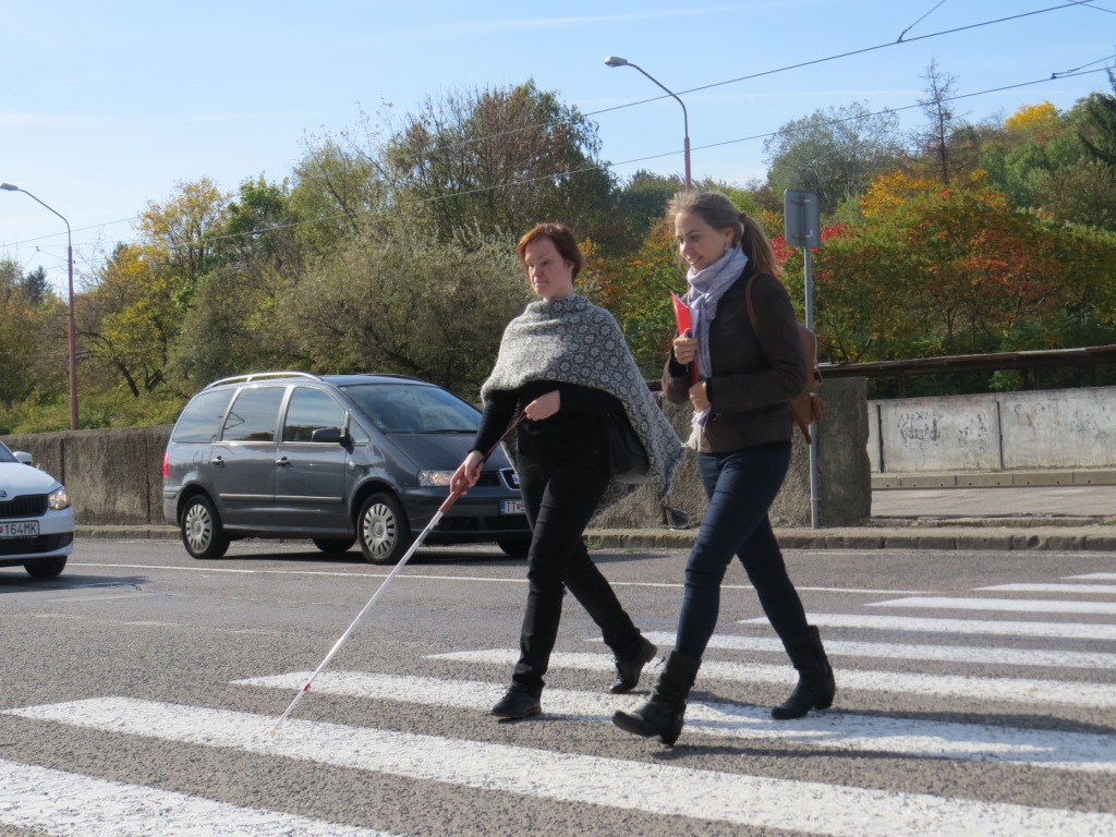 Cez prechod cez cestu kráčajú nevidiaci človek s bielou palicou v doprovode asistenta